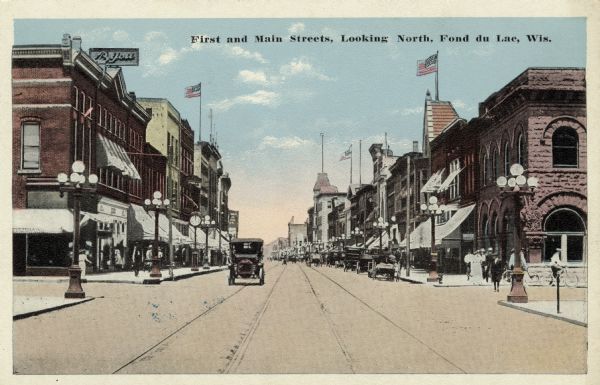View down middle of street towards the intersection of First and Main Streets. Automobiles, wagons and bicycles are on the street, and street lamps are along the sidewalks. Flags are flying from flagpoles. Caption reads: "First and Main Streets, Looking North, Fond du Lac, Wis."