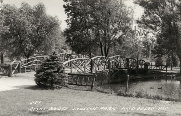 View of the rustic bridge in Lakeside Park featuring railings made of branches. Caption reads: "Rustic Bridge, Lakeside Park, Fond du Lac, Wis."