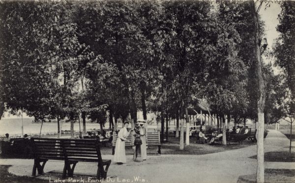 Scene at Lake Park. Two women are standing next to a drinking fountain. Several other people are sitting on the park benches. Caption reads: "Lake Park, Fond du Lac, Wis."