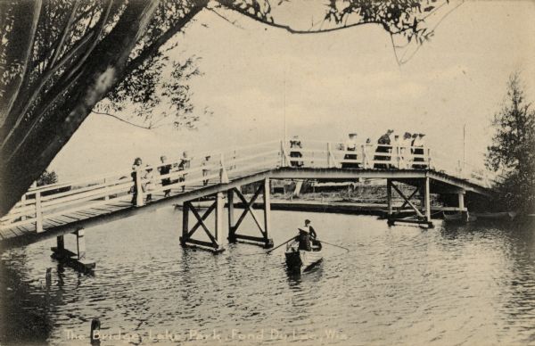 Elevated view of the Lake Park foot bridge, with women and children standing on it. A couple is in a rowboat below the bridge. Caption reads: "The Bridge, Lake Park, Fond du Lac, Wis."