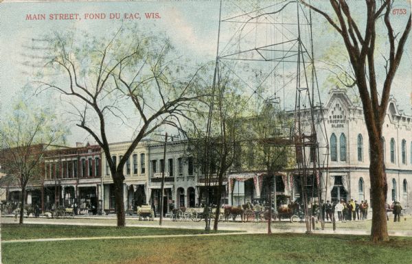 Hand-colored view across lawn towards Main Street, with buildings, horses, buggies and pedestrians. The base of a tower is between trees on the lawn in the foreground. Caption reads: "Main Street, Fond du Lac, Wis."