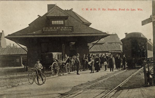 Photographic postcard view of a train on the tracks, several people on the platform, a boy standing with a bicycle, and a horse and carriage parked behind the platform. Caption reads: "C. M. & St. P. Railway Station, Fond du Lac, Wis."