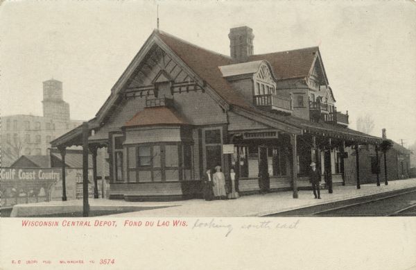 View of the Central Wisconsin Depot looking southeast. A few people are on the platform. There is a billboard for Gulf Coast Country across the street. Caption reads: "Wisconsin Central Depot, Fond du Lac, Wis." Penciled text reads: "looking south east."
