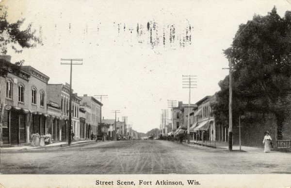 Photographic postcard view down the center of a unpaved street in downtown Fort Atkinson. Horse-drawn vehicles are at the curbs. Caption reads: "Street Scene, Fort Atkinson, Wis."