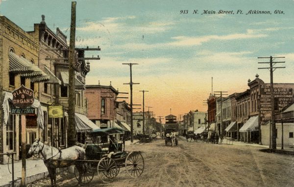 Hand-colored scene of north Main Street lined with businesses. There are horse-drawn vehicles in the unpaved street. Caption reads: "N. Main Street, Ft. Atkinson, Wis."