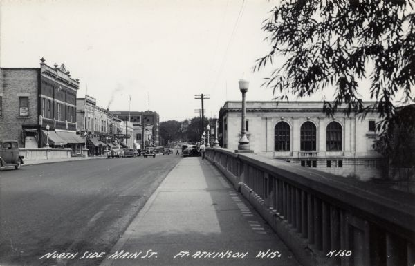 Black and white photographic view of Main Street from the vantage point of a bridge. Many automobiles are parked along the curbs, and the street is lined with lampposts and businesses. Caption reads: "North Side Main St., Ft. Atkinson, Wis."
