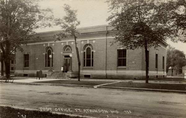 Photographic postcard view across street towards the post office, with a girl standing at the top of the steps at the entrance. A bicycle is parked at the curb. Caption reads: "Post Office, Ft. Atkinson, Wis."