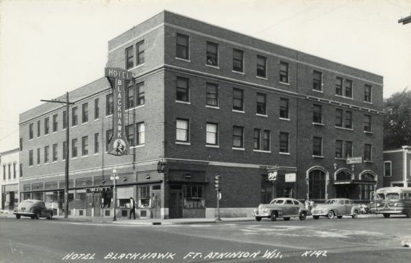Black and white photographic postcard of the Hotel Blackhawk, a large brick building on a corner in the downtown area. Automobiles and buses are at the intersection. On the far right is a sign for the Blackhawk Tavern. Caption reads: "Hotel Blackhawk, Ft. Atkinson, Wis."