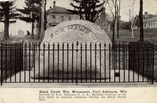 A granite stone behind a low fence commemorating the Black Hawk War. Text on front of card reads: "Black Hawk War Monument Erected by Fort Atkinson D.A.R., marking position of the fort held by General Atkinson during the Black Hawk War."