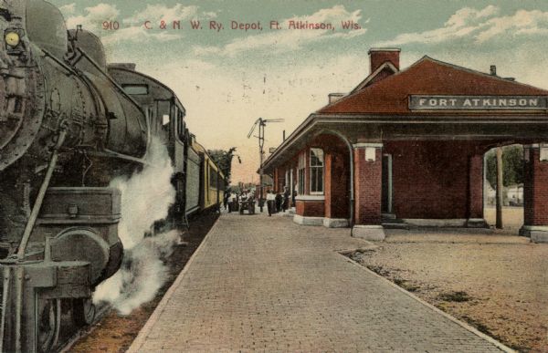 Hand-colored view of the train depot with a passenger train on the tracks and passengers on the platform. Caption reads: "C. & N.W. Railway Depot, Fort Atkinson, Wis."