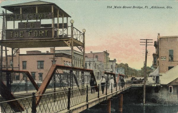 View of the Main Street Bridge with a large electric sign "Welcome to the Fort" on the left. Pedestrians are walking on the bridge. Caption reads: "Main Street Bridge, Ft. Atkinson, Wis."