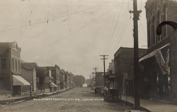 View of Main Street, with a barbershop on the left and a shoe store on the right. A buggy with two horses is parked at the curb. Caption reads: "Main Street, Fountain City, Wis., Looking South."