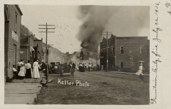 View down sidewalk and street towards the Fountain City fire of July 22, 1906. Smoke is billowing from a downtown building. Several onlookers are watching from the sidewalk and the street.