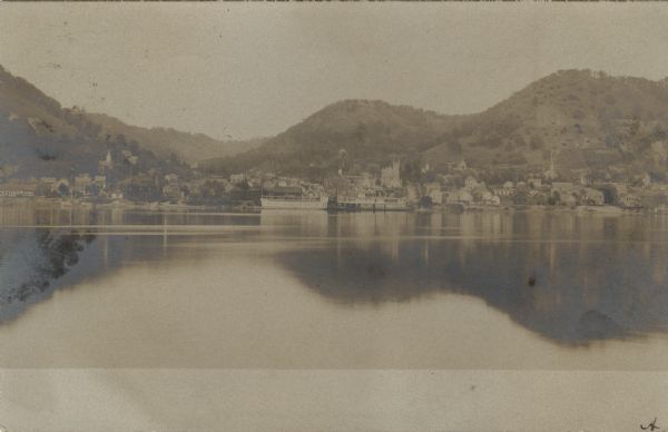 View of Fountain City from the Mississippi River. There is a steamer at the dock on the far shoreline. Hills in the background are reflected in the water.