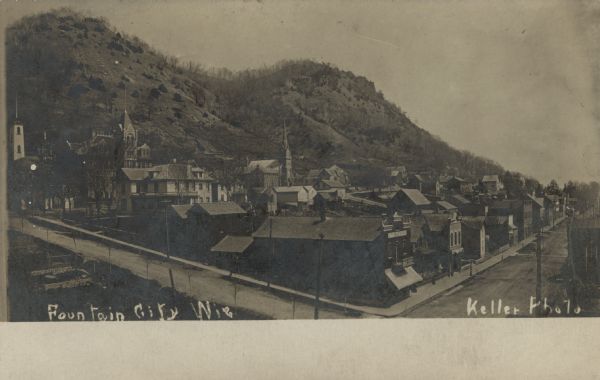 Elevated view of Fountain City, with a steep bluff in the background. In the foreground is a street corner with a drugstore and other storefronts. There are four churches, dwellings and other buildings. Caption reads: "Fountain City, Wis."