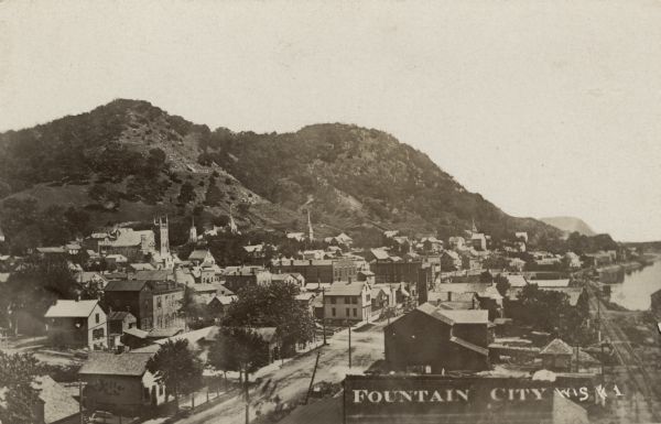 Elevated view of Fountain City, with the Mississippi River on the right, and hills in the background on the left. Caption reads: "Fountain City, Wis."