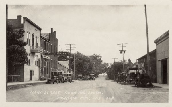 View down Main Street, with the Eagle Hotel on the left. Automobiles and trucks are parked along the curbs. Caption reads: "Main Street — Looking South, Fountain City, Wis."