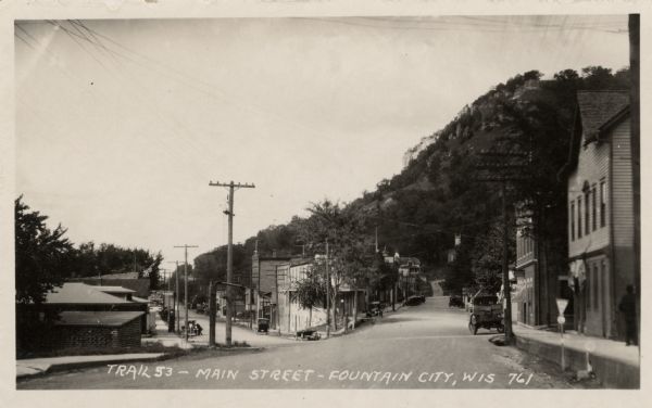 Intersection of Trail 53 and Main Street, lined with buildings. A road sign is in the foreground, and rucks and automobiles are parked at the curb. A steep bluff towers in the background. Caption reads: "Trail 53 — Main Street — Fountain City, Wis."
