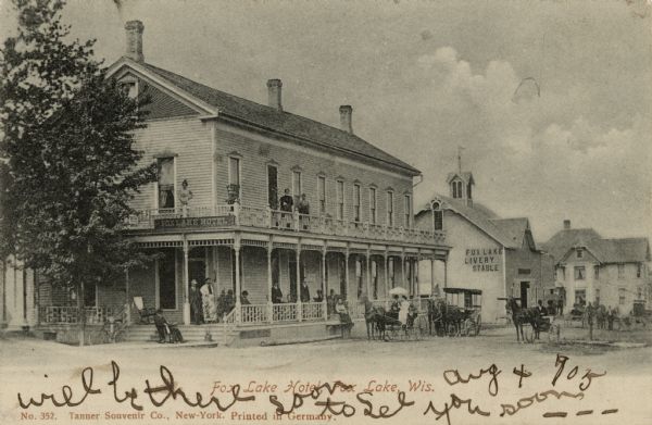 Exterior view of the Fox Lake Hotel, with several guests on the porch and balcony. Horses and buggies are in the street. The Fox Lake Livery Stable is next door. Caption reads: "Fox Lake Hotel, Fox Lake, Wis."