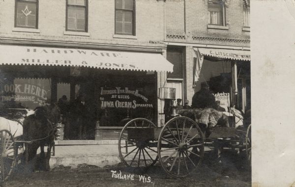View of the storefront of Miller & Jones Hardware, with two men standing at the entrance. Horses and carts are parked in the street. A Masonic symbol is in a second floor window. Caption reads: "Fox Lake, Wis."