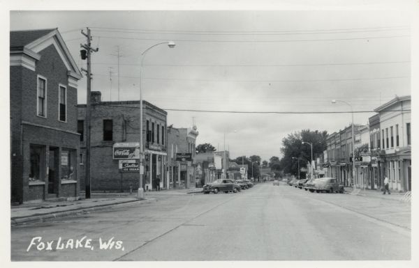 View down center of a city street, which is lined with bars, restaurants and other businesses. Automobiles are parked at an angle at the curbs. Caption reads: "Fox Lake, Wis."