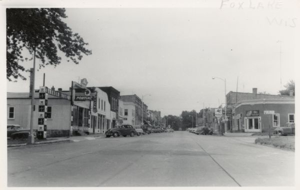 Photographic postcard view down center of street towards a commercial district. There are car dealerships, gas stations and other businesses, with automobiles parked at an angle at the curbs.