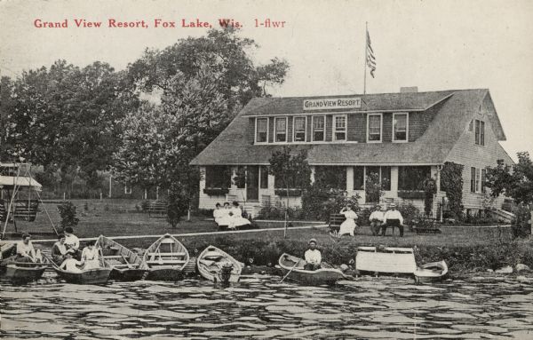 View across water towards the Grand View Resort on the shores of Fox Lake. Several people are sitting in rowboats lined up at the shoreline. Other guests are sitting on benches on the lawn. Caption reads: "Grand View Resort, Fox Lake, Wis."