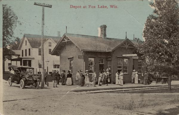 View across railroad tracks towards the railroad depot, with several passengers standing on the platform. An automobile is parked at the curb. Caption reads: "Depot at Fox Lake, Wis."