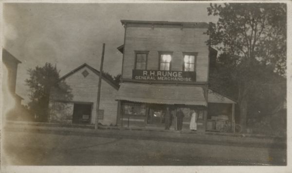 Exterior view of R.H. Runge's General Merchandise store. A woman and two men are standing near the entrance.

Penciled-in note on reverse reads: "Burned 6:30am Jan 10 1919"