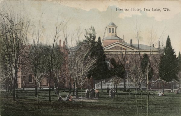 Slightly elevated view toward the Peerless Hotel, mostly obscured by trees. A group of women are in the park in the foreground. Caption reads: "Peerless Hotel, Fox Lake, Wis."