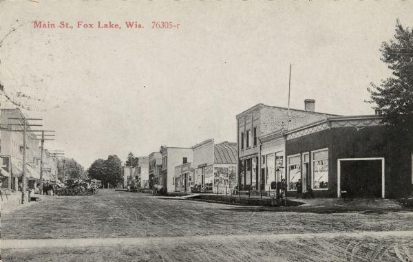 View down Main Street towards businesses. Several horse-drawn vehicles are along the curb on the left. Caption reads: "Main St., Fox Lake, Wis."