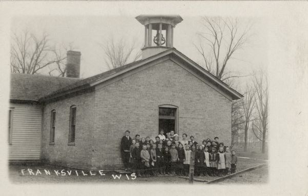 Students and teachers posing for a group portrait at the entrance of their school. There is a bell tower on the roof. Caption reads: "Franksville, Wis."