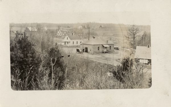 View from hill of Frederic, with a creamery in the center. Three men are standing near the doorway of the creamery, and an automobile is parked in front. Houses and farms surround the creamery.