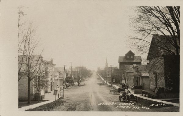 View down street with businesses, dwellings and a church. An automobile and a horse-drawn vehicle are parked on the right. Caption reads: "Street Scene, Fredonia, Wis."
