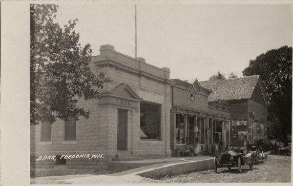 View down street towards the State Bank of Fredonia, and the general store next door. An automobile is parked parked along the curb. Caption reads: "Bank, Fredonia, Wis."