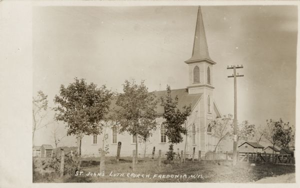 View down road towards the wooden church with a steeple. This church is located on the west side of the city on the north side of the road. Caption reads: "St. John's Lutheran Church, Fredonia, Wis."