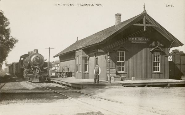 View across railroad tracks towards the Fredonia Depot. There is a train on the tracks, and a man is sitting on the front of the locomotive. A man is standing on the platform, and on the right, underneath the sign for Fredonia is a sign that reads: "Wells Fargo & Co. Express." Caption reads: "R.R. Depot, Fredonia, Wis."
