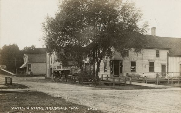 View across road towards the hotel next to the railroad tracks. A horse and buggy is parked nearby. There is a storefront on the block in the background. Caption reads: "Hotel & Store, Fredonia, Wis."
