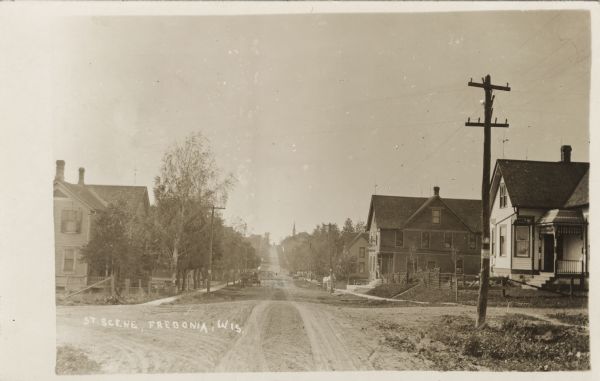View down unpaved street towards an intersection in a residential neighborhood. Horses and buggies are down the road near houses. There is a church at the top of the hill in the background. Caption reads: "St. Scene, Fredonia, Wis."