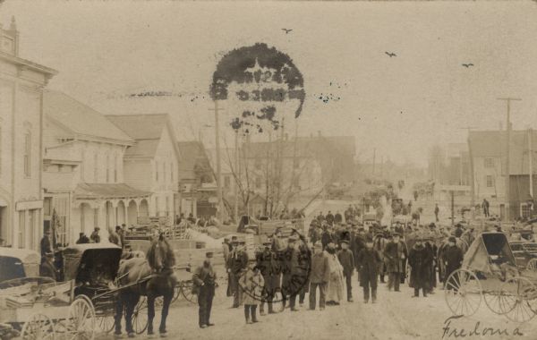 Elevated view of a busy street scene in downtown Fredonia. A crowd of people are standing among horse-drawn buggies.