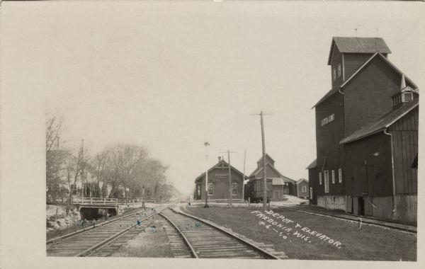 View down the railroad tracks, with the depot and grain elevator on the right. A bridge is crossing a stream on the left. Caption reads: "Depot & Elevator, Fredonia, Wis."