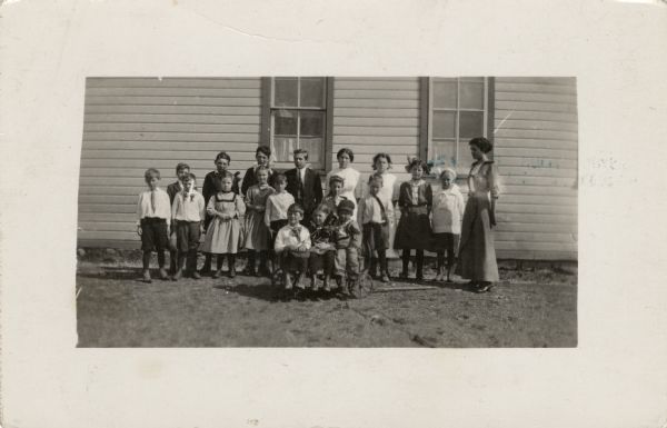A class of students ranging in age from 7 to 17 (?) and their teacher posing for a group portrait at the side of their school building. Three children are sitting in a wagon in front of the group.