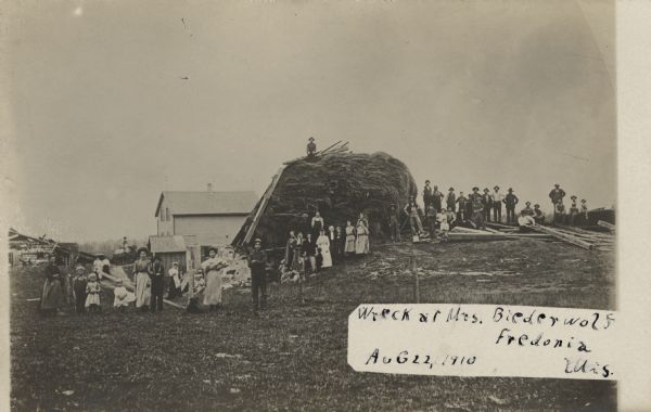 A crowd of people is posing near a haystack surrounded by debris. Caption reads: "Wreck at Mrs. Biederwolf, Fredonia, Wis."