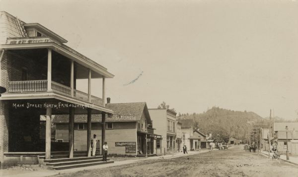 View down north Main Street. The Hotel Wrchota is on the left. Horse-drawn vehicles and automobiles are further down the street. A tree-covered hill is in the background.