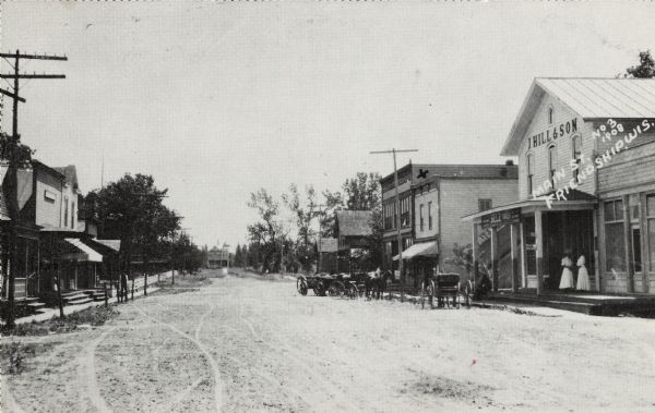View of Main Street in Friendship. The J.Hill & Son General Store is on the right, with two women at the entrance. Horse-drawn vehicles are in the street.