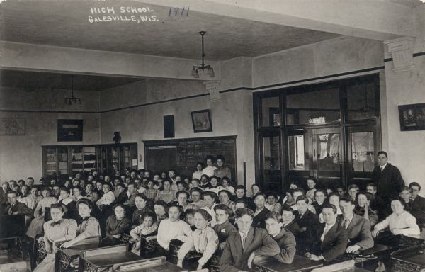 High school students are assembled in a classroom. Two students are sitting at every desk. Teachers are standing in the background.