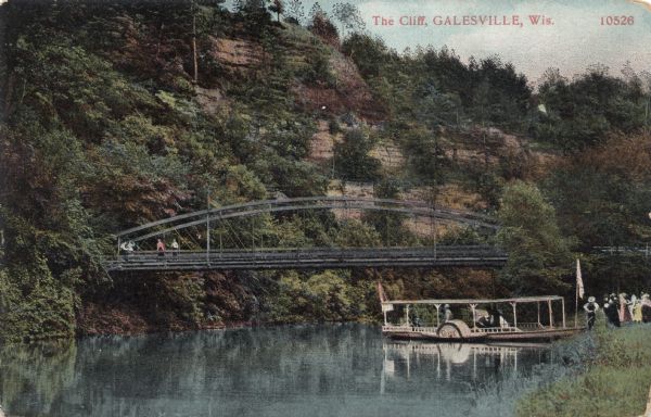View of a cliff overlooking a river. A group of people is gathered on the bank waiting to board an excursion boat. There is a bridge in the background.