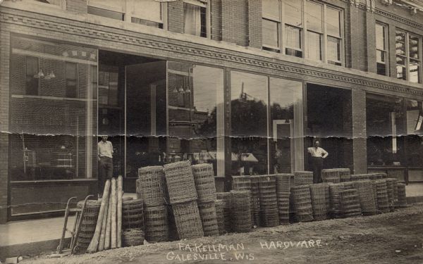 Exterior view of a hardware store, with two men standing outside. Rolled up fencing is at the curb.
