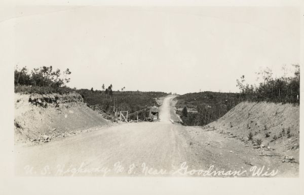 View down a dirt road stretching over a rolling hill. The land around it is clear of trees. Machinery is on the side of the road. Caption reads: "U.S. Hwy 8 near Goodman, Wis."