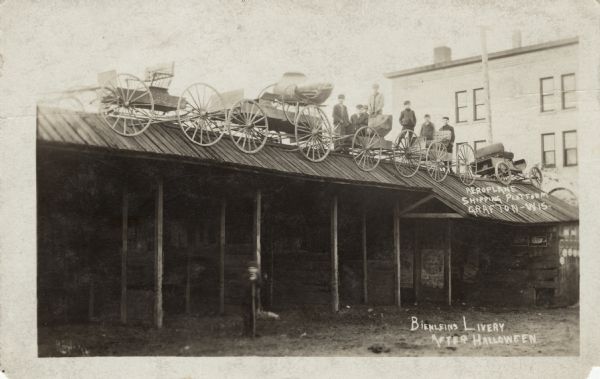 View of the livery stable, with a row of five buggies parked on the roof after Halloween. Seven boys are standing among the buggies on the roof, and another boy is standing in the yard. Text written on back reads, in part: "Aeroplane Shipping Platform." Caption reads: "Bienleins Livery After Halloween."